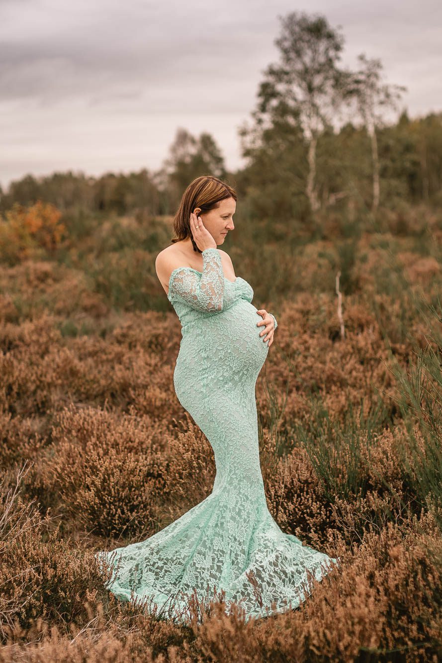 Schwangere stehend in einem schönen Kleid in der Heide, dabei hält sie ihren Bauch