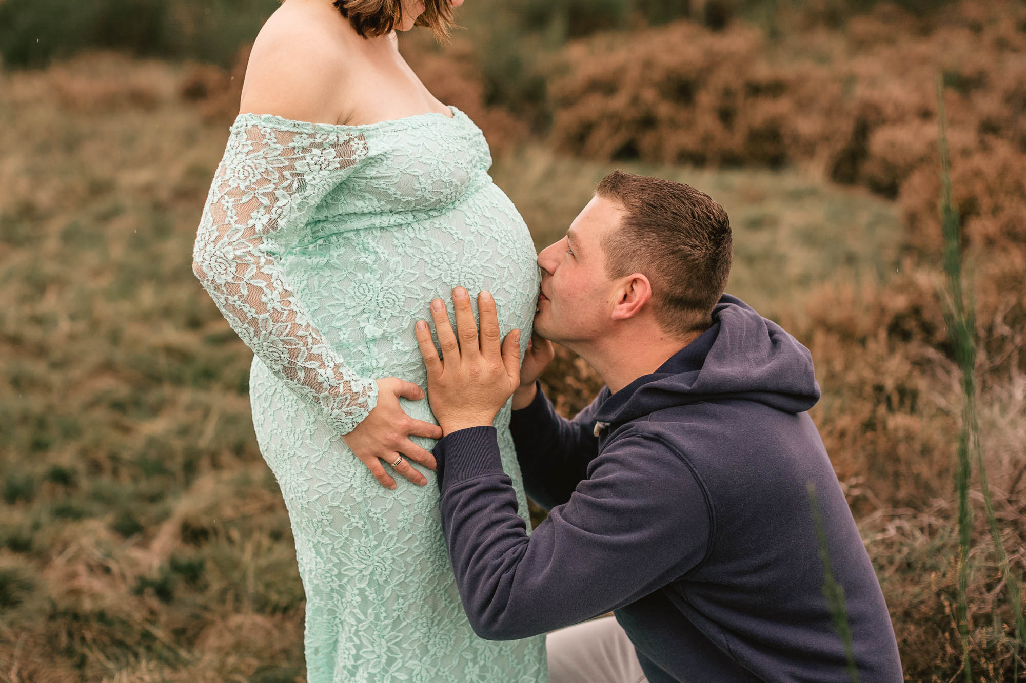 werdender Vater der den Babybauch seiner Frau hält und küsst vor ihr kniend während eines Schwangerschaftsshooting