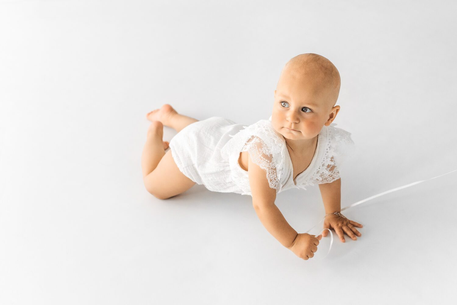 Baby welches im Fotostudio krabbelt und in Richtung Fensterlicht schaut, dabei ist es mit einem schönen weißen Romper bekleidet