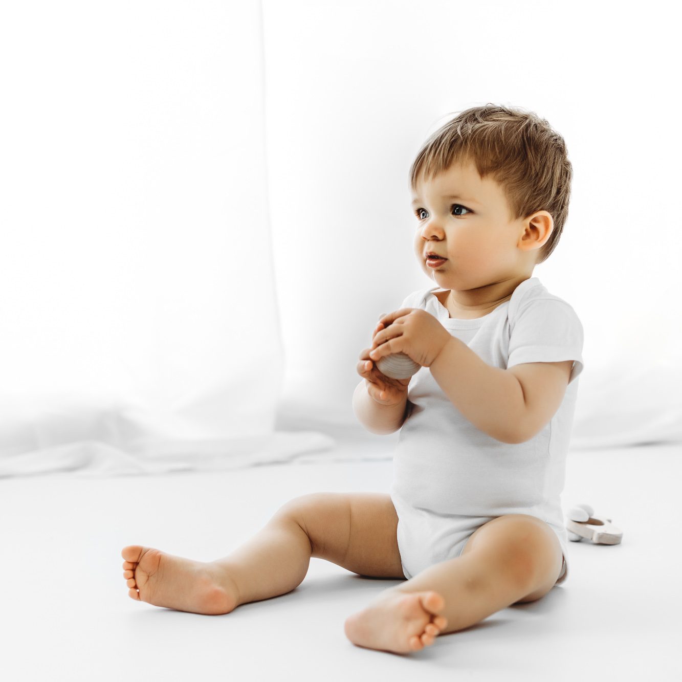 Baby Junge mit einem Holzspielzeug in der Hand im Fotostudio mit einem weißen Body sitzend