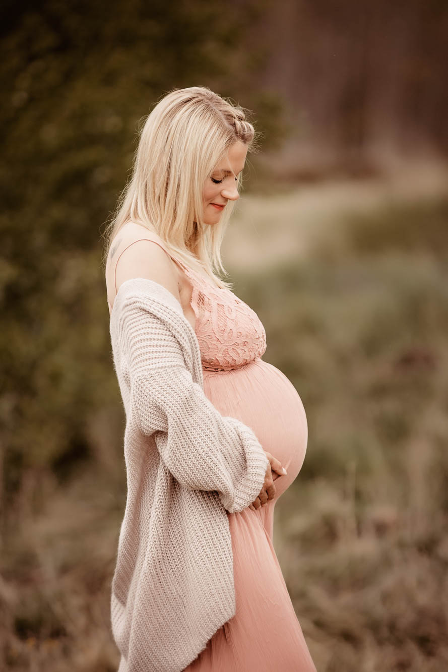 seitliches Profilbild einer schwangeren blonden Frau in einem schönen Kleid stehend schaut sie liebevoll auf ihren Babybauch
