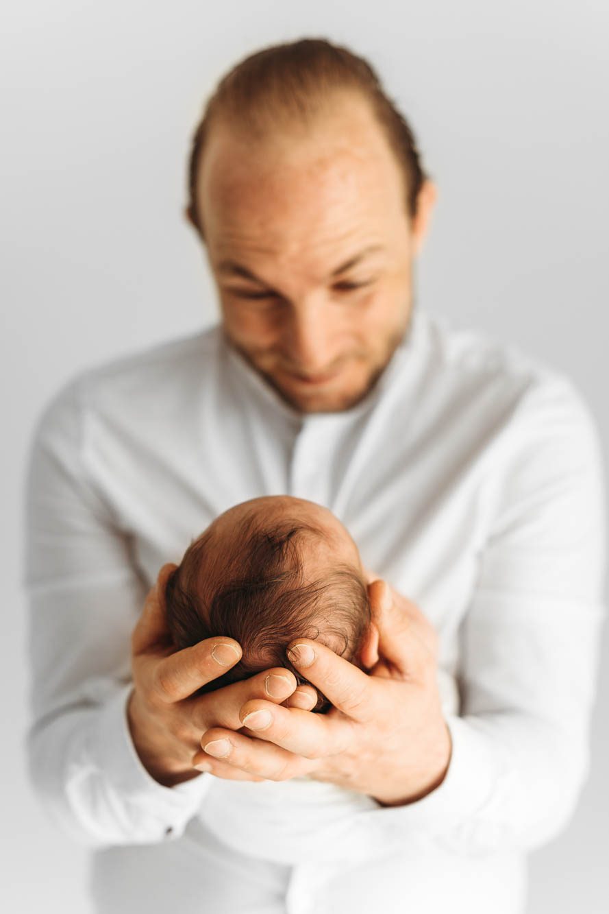 Vater der freudig sein Kind in den Händen hält, bzw. den Kopf seines neugeborenen Babies und dabei glücklich lächelt