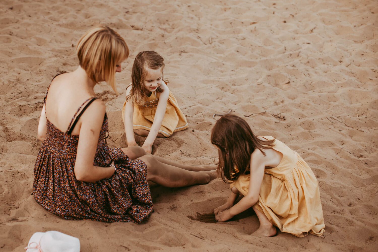 Familie im Sand sitzend und die beiden Töchter buddeln die Füße der Mutter ein