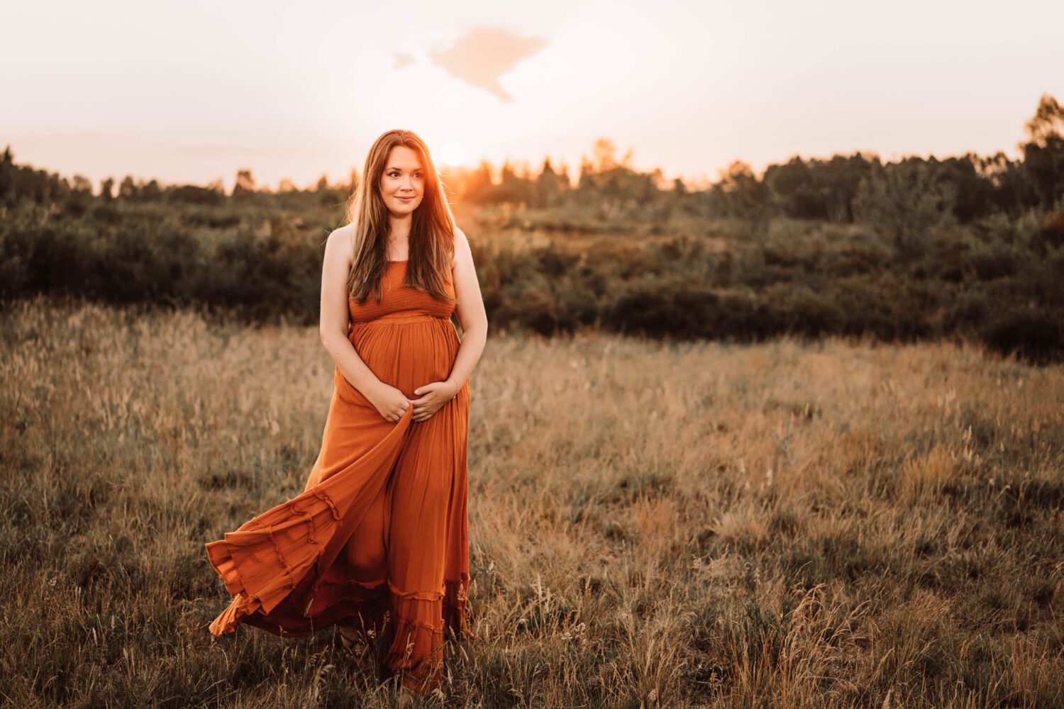 Schwangere im schönen langen Kleid strahlend und den Babybauch haltend läuft sie über eine Wiese im Abendlicht