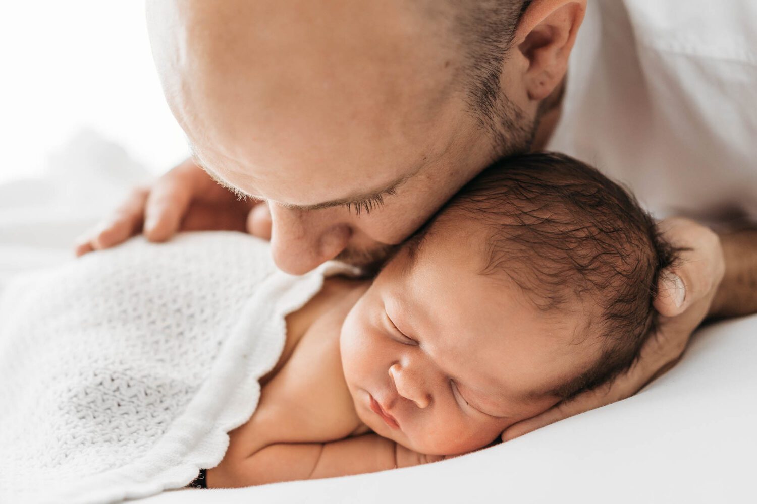 Neugeborenes Baby welches auf dem Bauch liegt mit dem Kopf in der Hand vom Papa, dieser Kuschelt sein Kopf ans das Baby