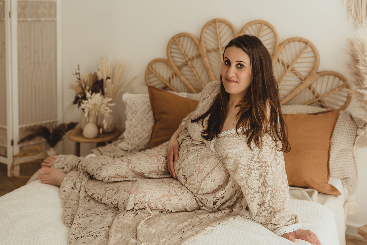 schwangere Frau auf einem Bett sitzend und dabei hält sie ihren Bauch und hat ein schönes Kleid an