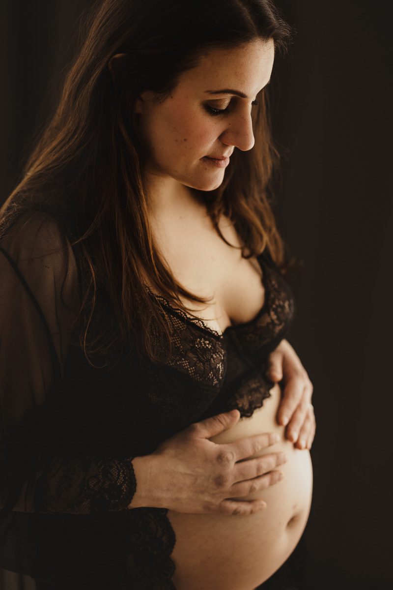 Schwangere vor einem dunklen Hintergrund mit hübscher schwarzer Unterwäsche seitlich im Porträt fotografiert wie sie ihren Bauch hält