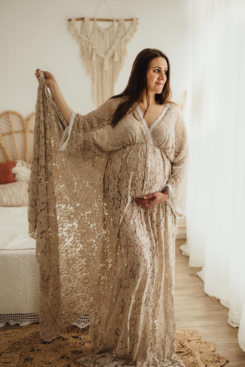 Schwangere stehend in einem Boho Kleid schaut sie verträumt Richtung Fenster und hält ihren Bauch dabei