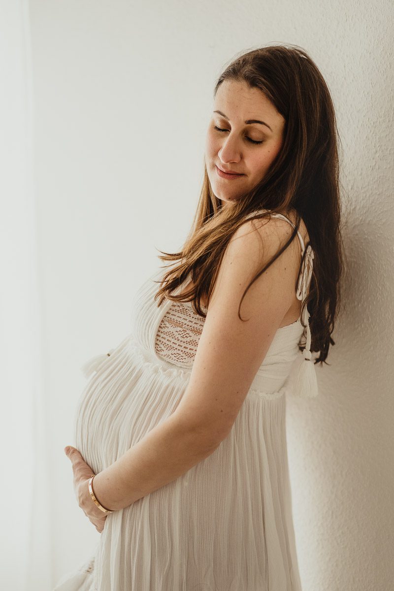 Schwangere in einem schönen weißen Kleid im Profil von der Seite fotografiert, sie hält dabei ihre Augen geschlossen und hält ihren Babybauch