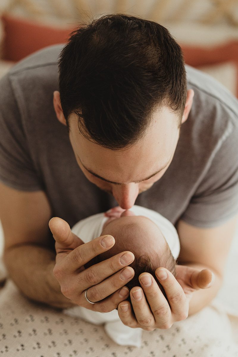 junger Vater von vorn fotografiert wie er sein neugeborenes Baby näselt, dabei liegt es mit seinem Köpfchen in seinen Händen
