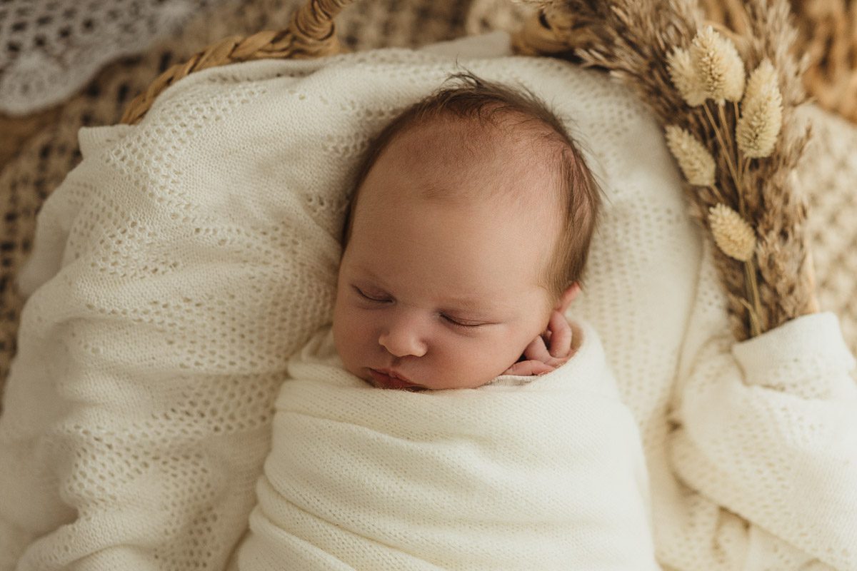 neugeborenes Baby liegt gepuckt neben Trockenblumen in einem Korb auf einer schönen cremefarbenen Decke dabei schläft es